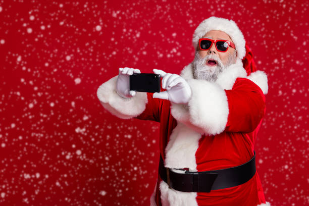 istock Portrait of attractive bearded amazed funny Santa using device taking making selfie vlog isolated over bright red color background 2184117423