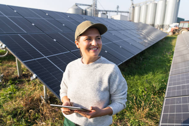 istock Female farmer wearing work uniform holds digital tablet in her hands stands next to solar panels. Modern agricultural farm using solar panels. Agricultural silos in the background. 2184122159