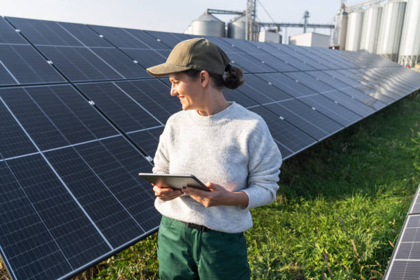 istock Female farmer wearing work uniform holds digital tablet in her hands stands next to solar panels. Modern agricultural farm using solar panels. Agricultural silos in the background. 2184122711