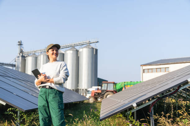 istock Female farmer wearing work uniform holds digital tablet in her hands stands next to solar panels. Modern agricultural farm using solar panels. Agricultural silos in the background. 2184122779