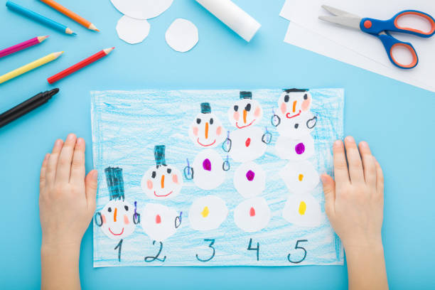 istock Little child hands creating and decorating snowman shapes and learning numbers on paper on light blue table background. Pastel color. Closeup. Point of view shot. Toddler development. Top down view. 2184126492