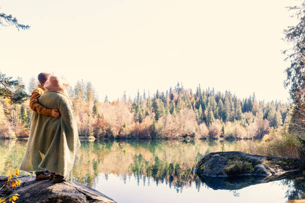 A couple shares a warm embrace wrapped in a cozy blanket by a serene lake, surrounded by autumn foliage. A peaceful moment amidst nature’s beauty.