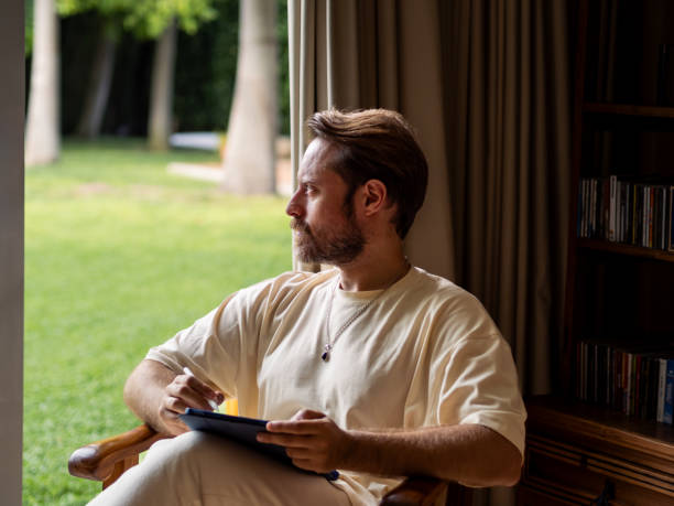 istock Man with tablet reflecting while seated indoors, enjoying a garden view 2184150792