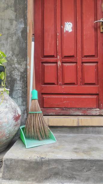 A wooden broom propped up against a wall.  The wall is two different colors.  The top half of the wall is solid chocolate brown.  The bottom half of the wall is bead board, and it has repeating parallel lines spaced a few inches apart.  The bottom section of the wall is a light shade of blue.  Propped up against the wall is a tan wooden broom.  The broom has angled bristles gathered with a dark thread.  There is white molding separating the two sections of the wall and at the floor, which is comprised of light-colored wood.