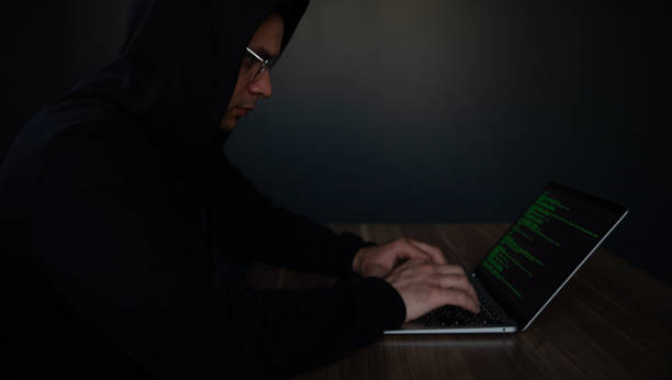istock A man in a black hoodie and glasses types on a laptop with green code on the screen. The image emphasizes cybersecurity, hacking, programming, or tech-related work in a dark, focused setting. 2184155002