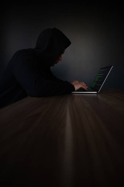 istock A hooded individual is focused on typing on a laptop with green code on the screen, sitting alone at a long wooden table in a dark, minimalist room, symbolizing hacking or cybersecurity themes. 2184155023