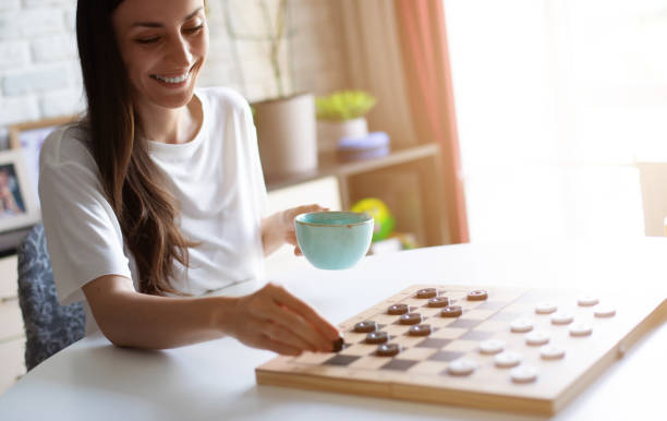 A smiling cute woman plays a checkers game at a white table while holding a teal mug. The scene is set in a cozy, sunlit room, conveying relaxation, joy, and a casual, playful atmosphere.