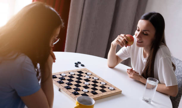 istock Two young girlfriends sit at a round table playing checkers. One is deep in thought, while the other smiles, holding an apple. A wooden board is in the center, with pieces in play. 2184165428