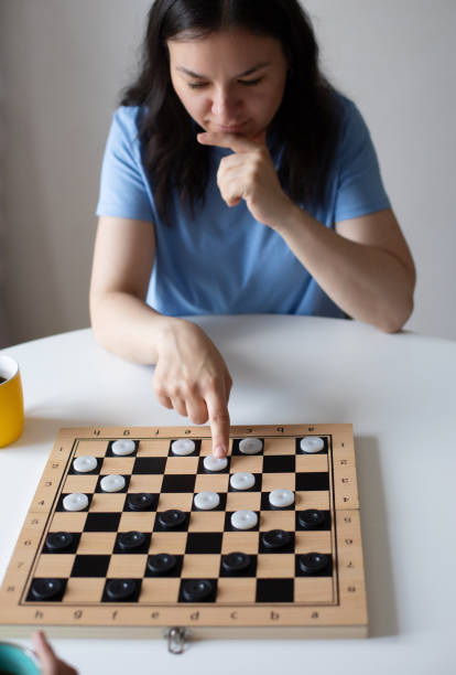 istock A focused woman in a blue shirt contemplates her next move while pointing at a piece on a checkerboard. The scene emphasizes strategy and thought during a friendly game in a casual setting. 2184165965