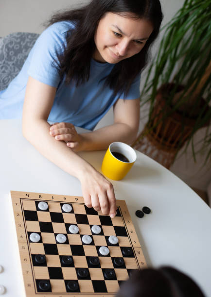 istock Woman in a blue shirt smiles while making a move in a checkers game at a white table. A yellow mug filled with coffee sits beside her. The scene is cozy and relaxed, set in a casual home environment 2184167227