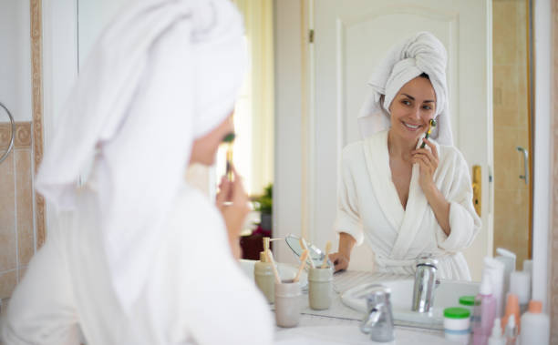 istock Woman in a bathrobe and towel on her head uses a jade roller on her face while smiling in front of a bathroom mirror. Skincare products are arranged on the counter, emphasizing self-care and beauty 2184170882