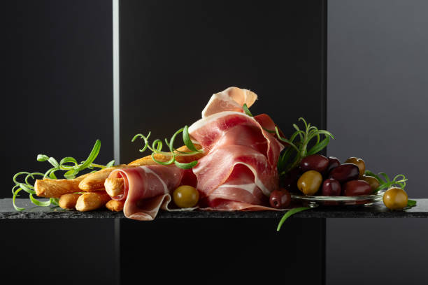 Prosciutto with bread sticks, olives, and rosemary on a black background.