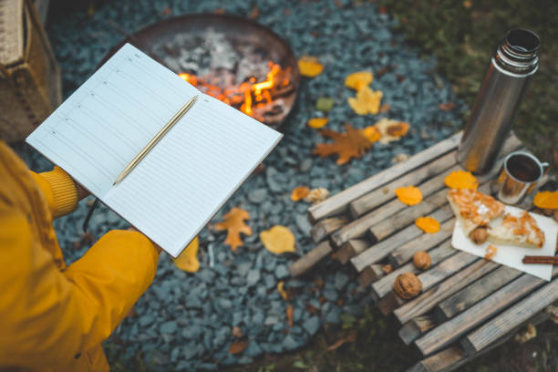 istock Person writing in a notebook next to a bonfire during autumn. Outdoor cozy setting with fallen leaves, snacks, hot drink, and the warmth of the fire, capturing the essence of a peaceful autumn day 2184173131