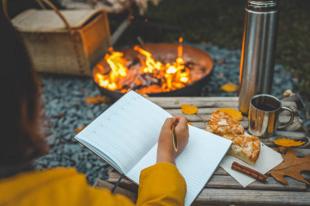 istock Person writing in a notebook next to a bonfire during autumn. Outdoor cozy setting with fallen leaves, snacks, hot drink, and the warmth of the fire, capturing the essence of a peaceful autumn day 2184173676