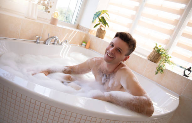 istock A cheerful man is relaxing in a bright bathroom, enjoying a bubble bath. The scene is warm, with sunlight streaming through the windows, plants, and bathroom products adding a cozy, inviting touch. 2184175432