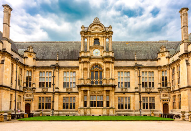 A pair of kids are positioned on a stone pathway in front of an ancient structure of All Souls College flanked by a green yard under a sunny sky, England
