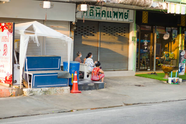 Three thai senior women with protective face masks are sitting in front of buildings with closed jalousies. Two are sitting on a small  stony bench. Scene is in street Senanikhom Road. One women is using a mobile phone. Women are wearing casual clothes. At right side is a small coffee shop. In front is a small sign.