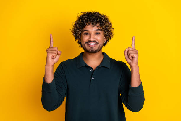 istock Young man in black shirt pointing upwards against bright yellow background, smiling confidently, with curly hair 2184176367