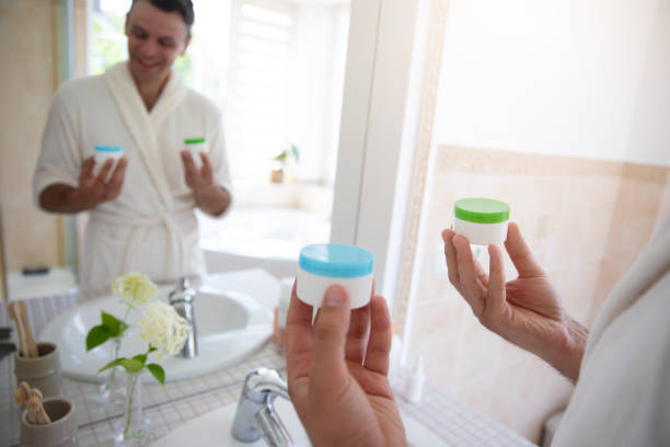 istock Young man in a white bathrobe stands in front of a bathroom mirror, smiling while comparing two skincare containers. The modern bathroom features a tiled countertop, plants, and natural lighting. 2184176675