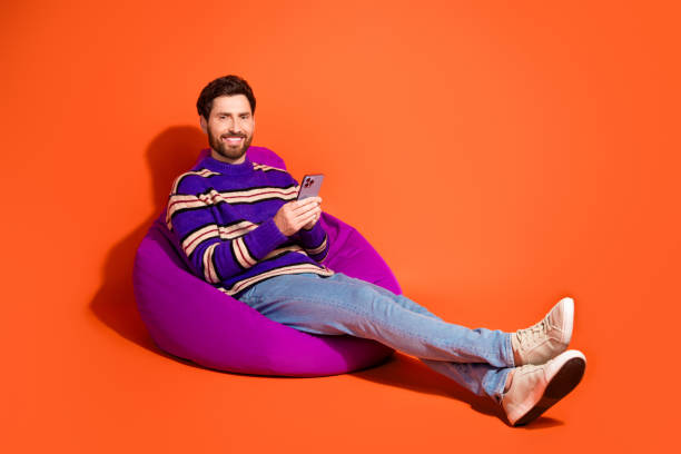 istock Young bearded man relaxing on a purple bean bag while using a smartphone over vibrant orange background 2184179534