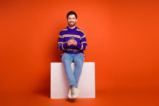 istock Full length photo of handsome young guy sit white cube hold device dressed stylish striped purple garment isolated on orange background 2184179841