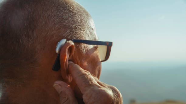 An elderly Caucasian man tunes his hearing device in nature, enhancing his auditory experience during an active day of hiking.