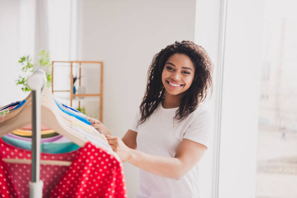 istock Smiling young woman browsing clothes rack with daylight streaming into modern living room apartment 2184183914