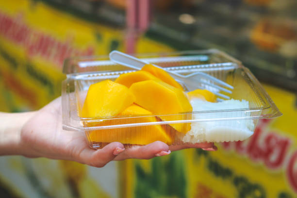 tourist eating fresh mango slices with sticky rice and coconut milk in a plastic container in front of a street market stall in Bangkok, Thailand