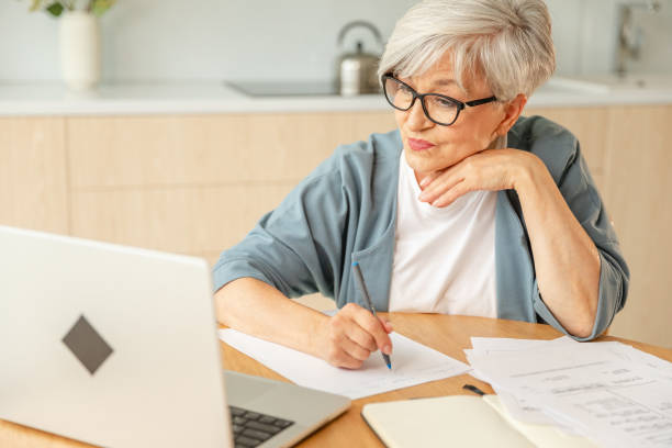 istock Middle aged senior woman using laptop computer writing notes at home. Focused mature old businesswoman work in home office making written records doing online work. Mature boss professional worker. 2184199968