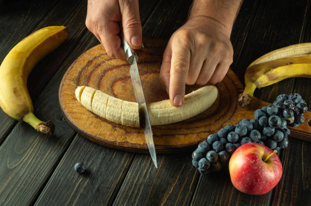istock Close-up of chef hands with knife cutting banana before preparing fruit dessert on kitchen table. Concept of preparing healthy dinner with fruits 2184205324