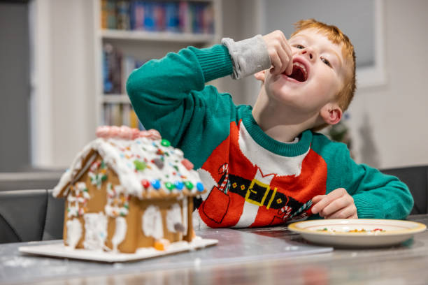 Little redhead boy making a gingerbread house for Christmas. He is in the kitchen and having fun while putting the icing and the candies.