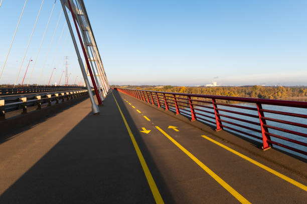 A straight view along the biking lane on the Pentele Bridge, featuring yellow directional arrows and red railings with autumn foliage and an industrial skyline in the distance under a clear blue sky.