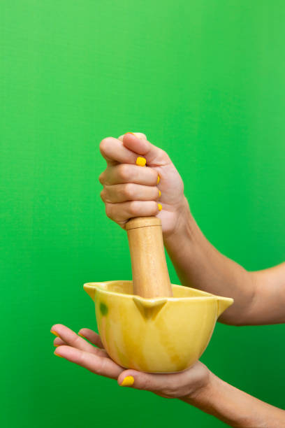 Hands of a woman in her forties holding a yellow ceramic mortar with her wooden mallet on a green background.