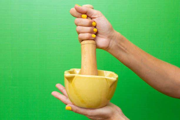 Hands of a woman in her forties holding a yellow ceramic mortar with her wooden mallet on a green background. Vertical photograph