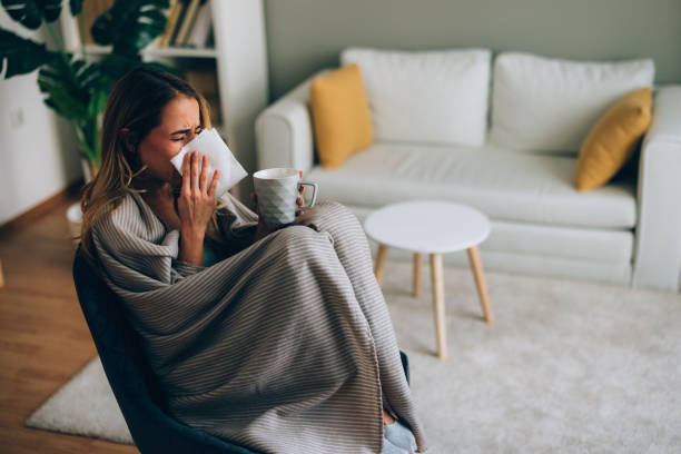 istock Young woman covered with blanket is sneezing and drinking a hot beverage while sitting on an armchair at home 2184212933