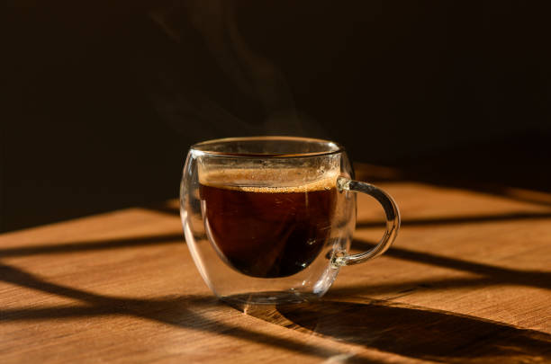 istock Warm cup of coffee steaming on a wooden table with dramatic sunlight filtering through window shadows 2184213301
