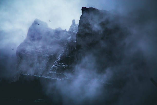 A misty mountain scene with towering dark cliffs partially obscured by thick fog. Four birds are silhouetted against the blue-gray sky, adding an eerie, mysterious atmosphere to the rugged landscape.