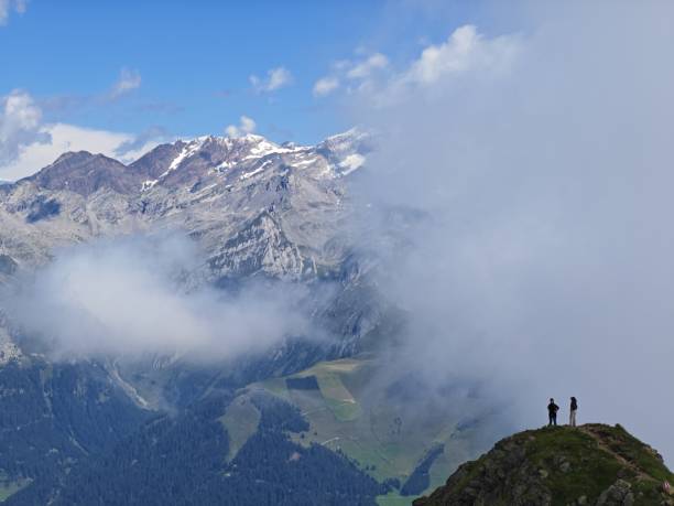 At the summit, two hikers take in a breathtaking view of the snow-capped mountains while clouds drift across the landscape, creating a serene atmosphere.