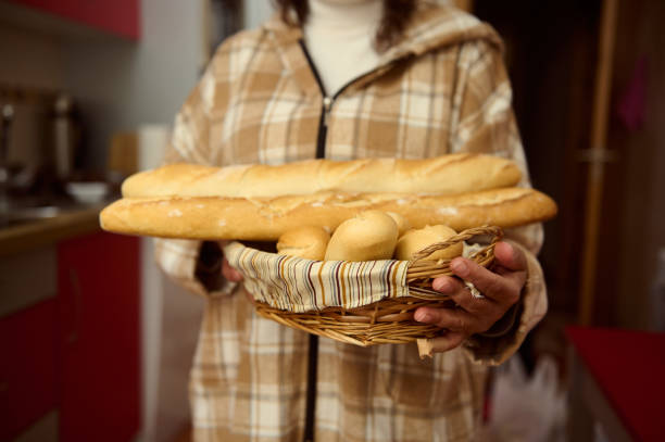 A person in a cozy kitchen holds a basket with two fresh baguettes and small bread rolls. The warm, inviting scene suggests a homemade and comforting environment, emphasizing artisanal baking.