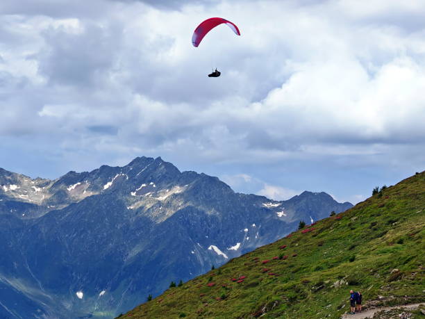 A paraglider gracefully glides above a stunning mountain landscape, with lush greenery on the slope and dramatic peaks in the background. Clouds add depth to the scene.