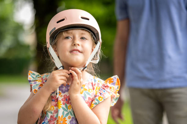 A cheerful young girl with a floral top fastens her protective helmet under adult supervision, preparing for a safe cycling adventure in a park setting.