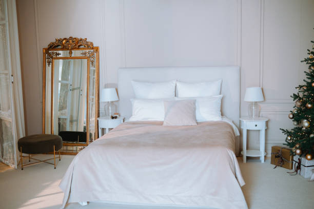 A warm and inviting bedroom features a beautifully made bed, soft pillows, and tasteful decor. A charming Christmas tree adds a festive touch to the serene atmosphere, perfect for relaxation.