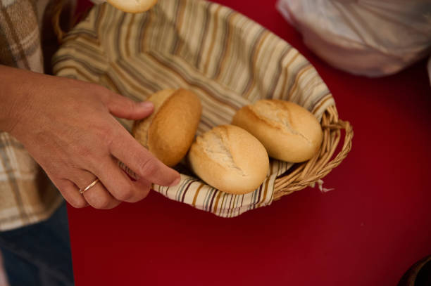 A close-up of a hand placing rustic bread rolls into a lined woven basket. The setting evokes a warm, homely atmosphere with emphasis on fresh, homemade bakery products.