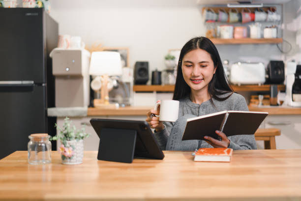 istock woman enjoys cozy moment at home, sipping coffee while reading notebook and using tablet. warm kitchen setting enhances relaxed atmosphere. 2184253882