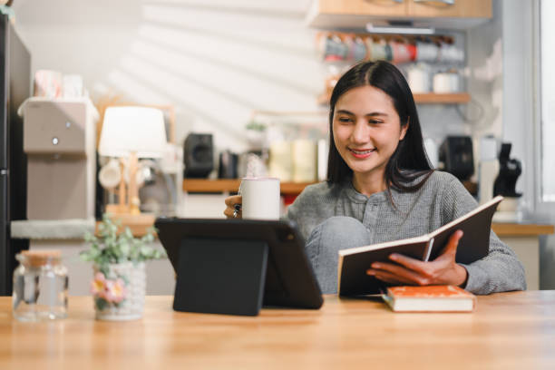 istock woman enjoys cozy moment in her kitchen, sipping drink while reading book and using tablet. warm atmosphere creates sense of relaxation and contentment. 2184253885