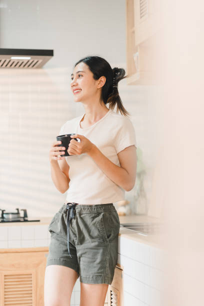 woman stands in bright kitchen, holding cup and smiling. She wears casual outfit, exuding relaxed and cheerful vibe. warm atmosphere enhances her joyful expression.