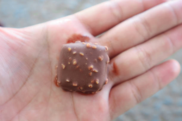 A close-up of a small, square chocolate with a hazelnut topping held in a person's hand. The chocolate has a glossy finish and the hazelnuts are visible on the surface.