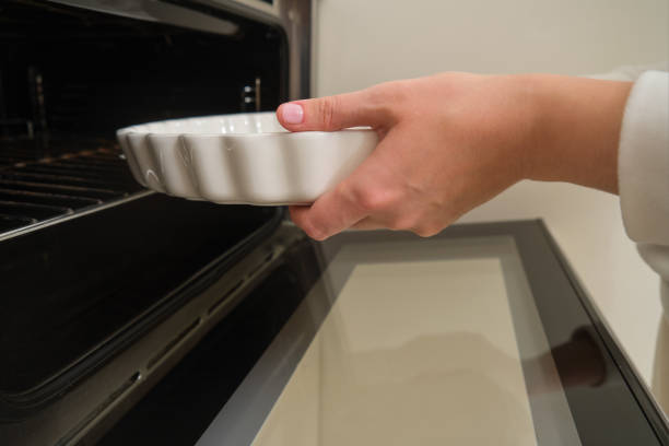 Hand placing white ceramic baking dish into open oven. Close-up food preparation concept for kitchen and baking content.