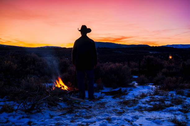 Cowboy Camp with Wrangler Man Tending to Fire in Mountain Setting - Rugged rancher outdoorsman at dusk with fire glowing tending to camp at high camp in mountains. Scenic dusk sky and mountain backdrop with camp at lower elevation among sagebrush meadows.