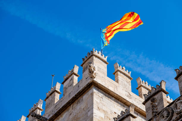 Detail of Lonja de la Seda tower in Valencia with spires, stone figure, waving Valencian flag, and clear blue sky.